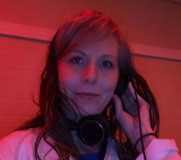 A woman with headphones holding one earpiece, smiling slightly in a room lit with red ambient light.