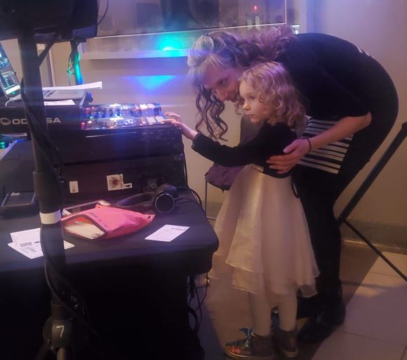 A woman and a young girl with curly hair interacting over a dj console at an indoor event.