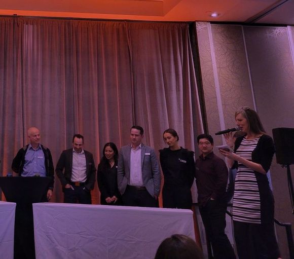 A group of professionals standing on stage, listening to a woman speaking into a microphone at a formal event.