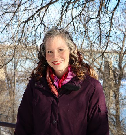 A woman with curly hair, smiling, wearing a burgundy jacket and a pink scarf, standing outdoors with bare trees and a river in the background.