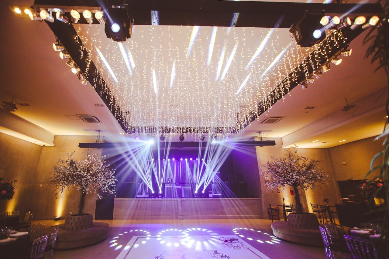 Elegant indoor event venue with purple lighting, a dance floor with decorative patterns, and a ceiling adorned with shimmering lights, surrounded by floral arrangements.
