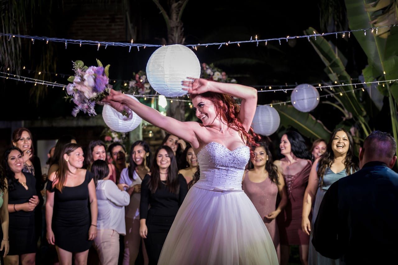 A bride joyfully throws her bouquet to a crowd of excited guests at an outdoor evening wedding reception.