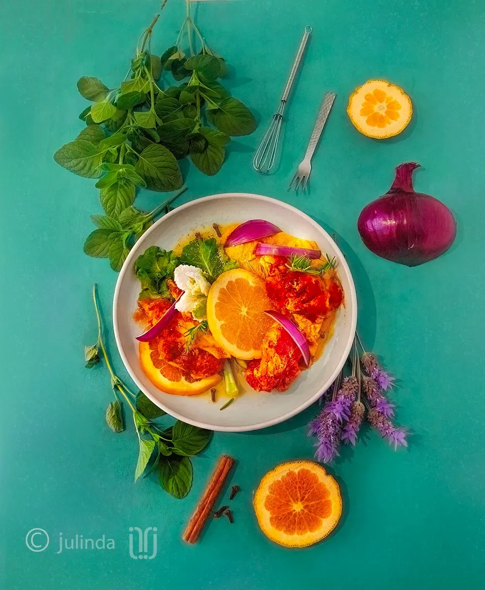 Overhead photography of a plate of organic chicken garnished with orange slices and red onion, with mint sprigs, a cinnamon stick, and a purple onion scattered around. Auckland food photographer.