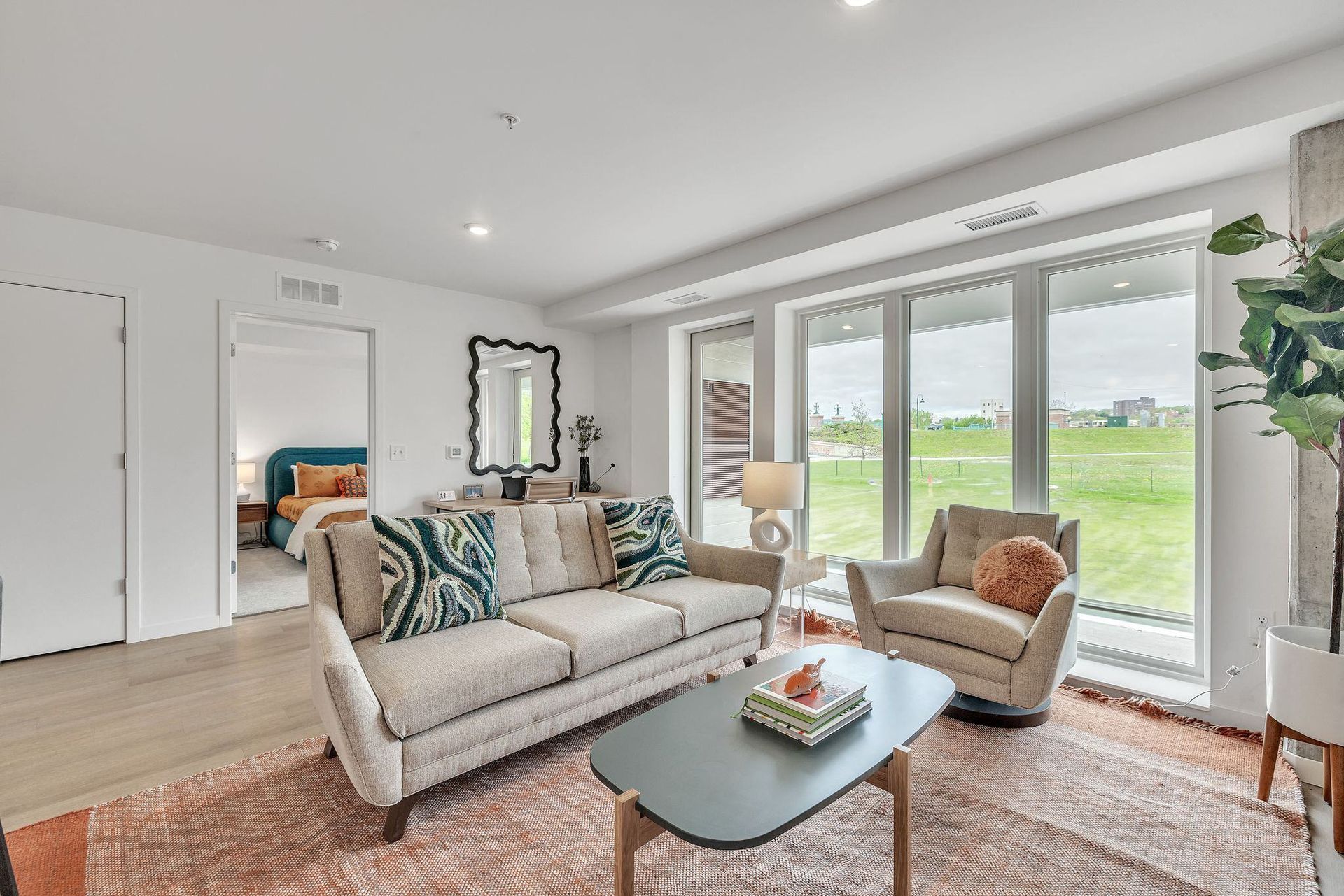 Bright living room with beige sofa, armchair, and large windows overlooking green space at Esox House in St. Paul, MN.