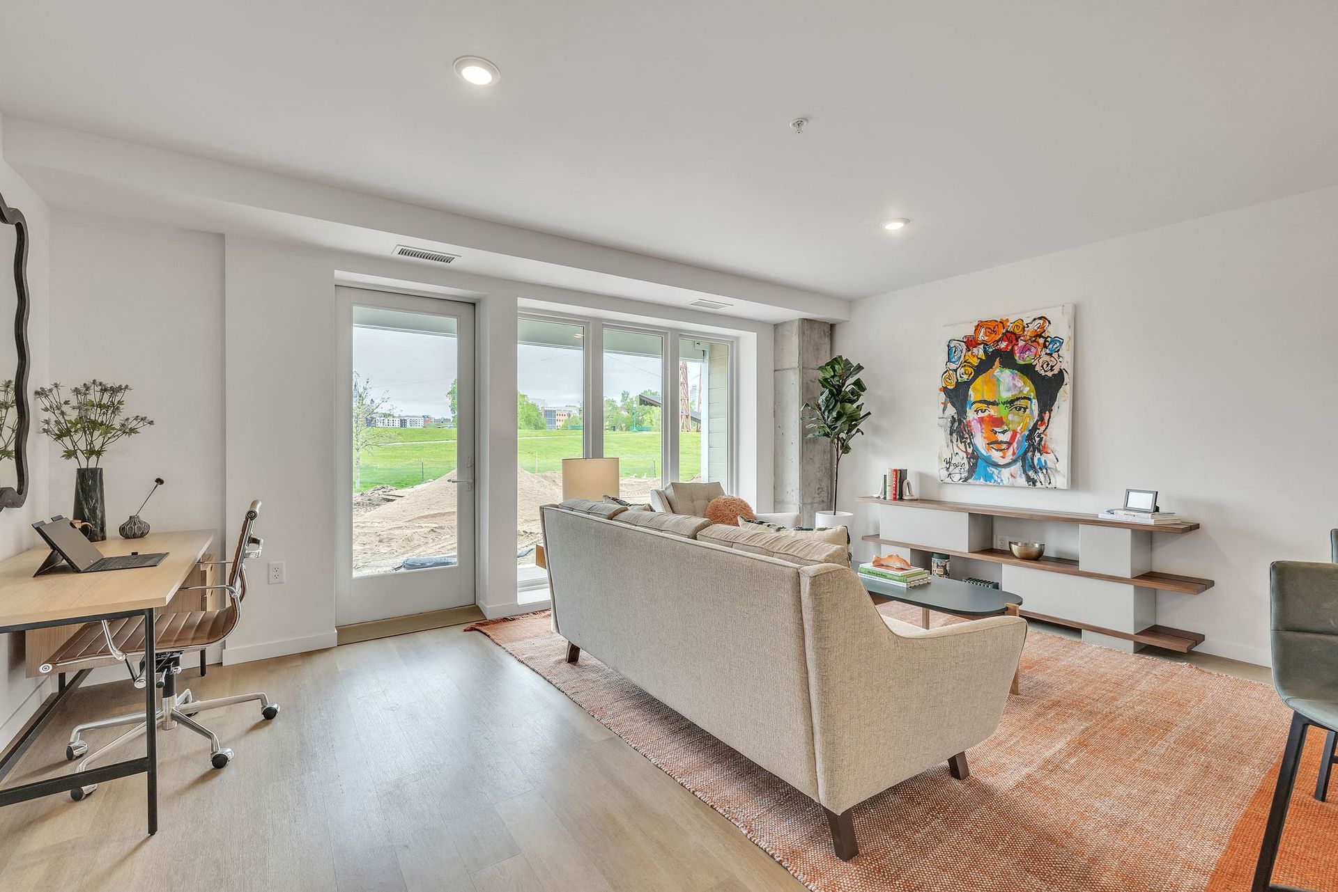 Bright living room with beige sofa, desk, wall art, and glass doors to a green outdoor view at Esox House in St. Paul, MN.