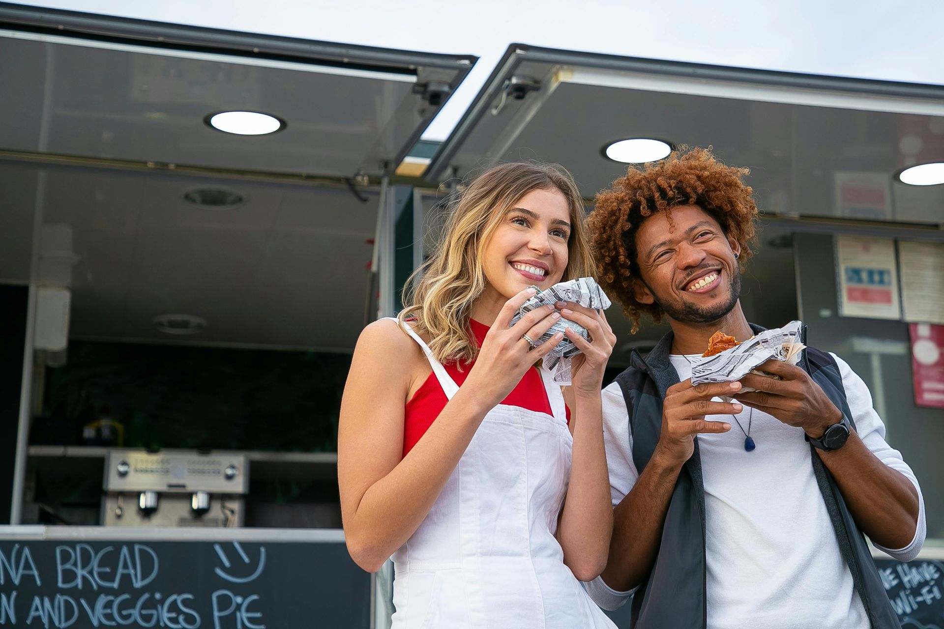 Two people stand outside a food truck eating wrapped food and smiling.