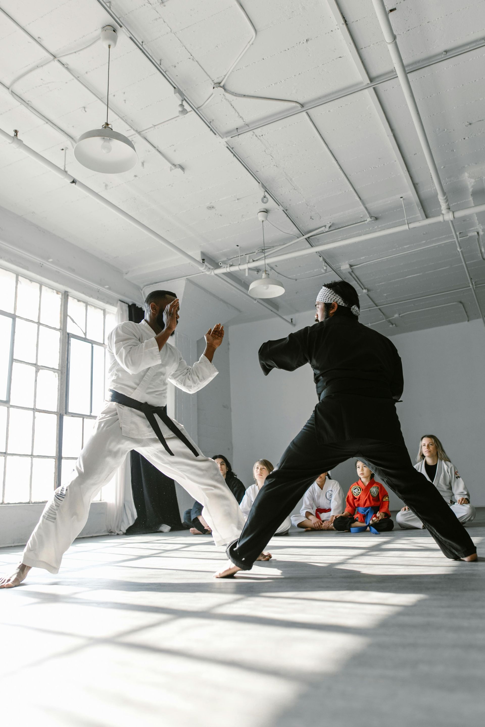 Two martial artists in gi spar in a sunlit studio, with other students sitting on the floor in the background.