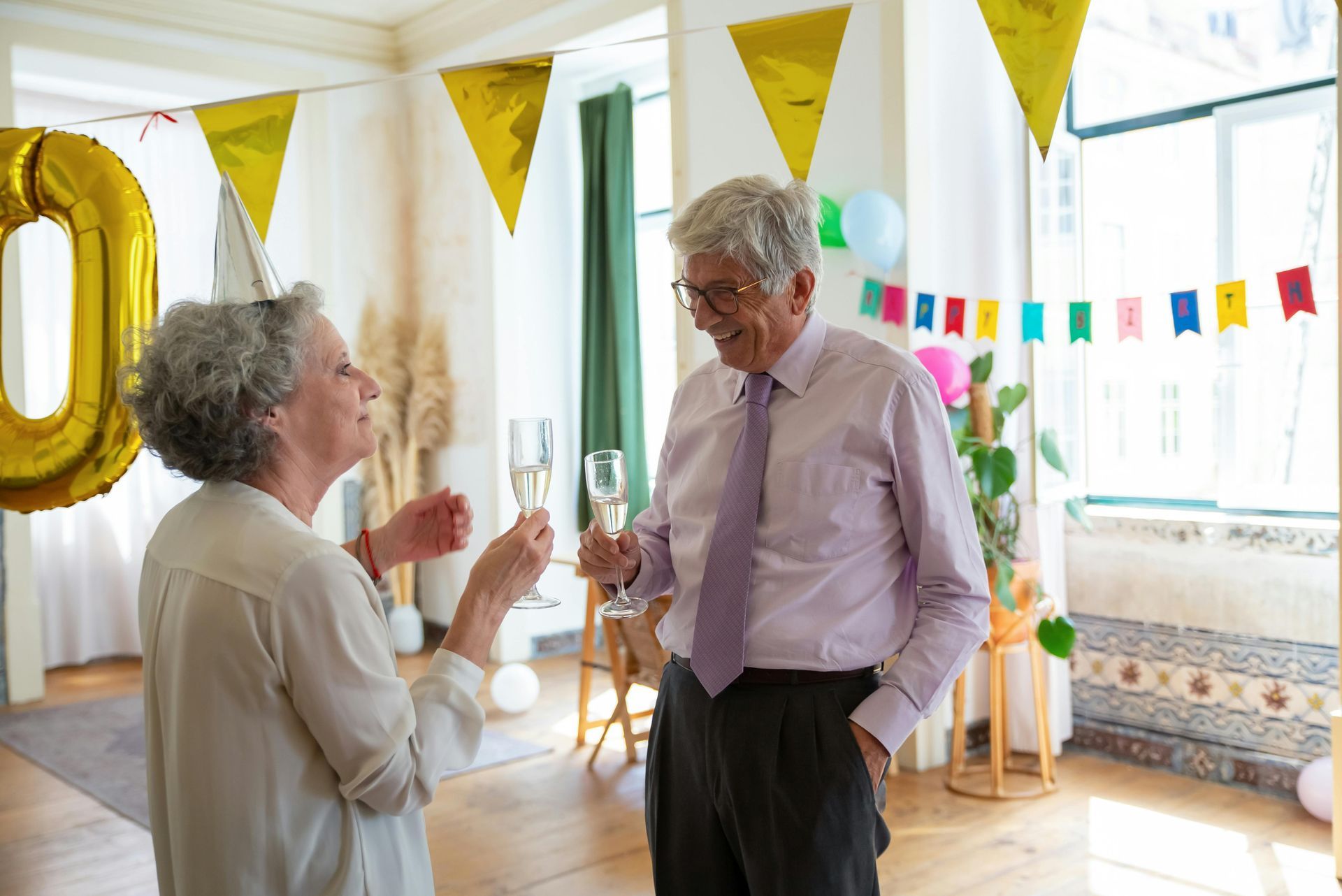 Two people in a decorated room hold champagne flutes and toast during a celebration under gold triangular banners.