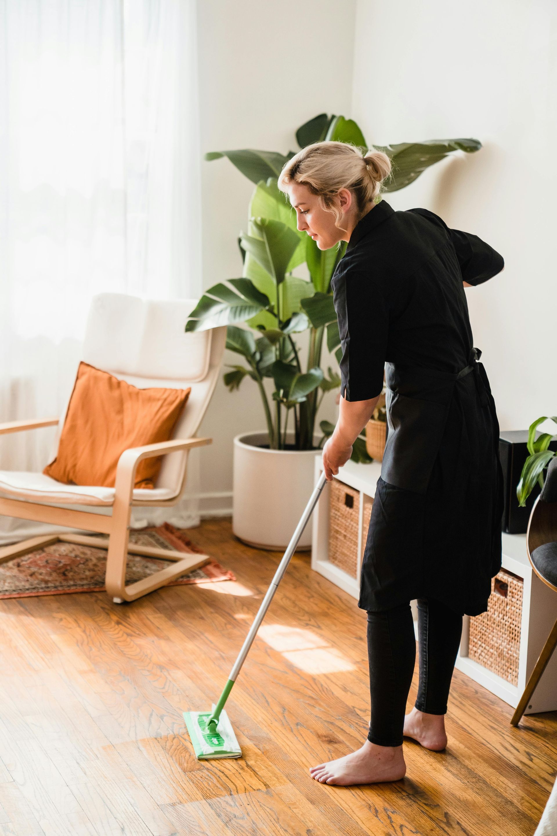 A person in black clothing mops a hardwood floor in a brightly lit room with a large potted plant and an armchair.