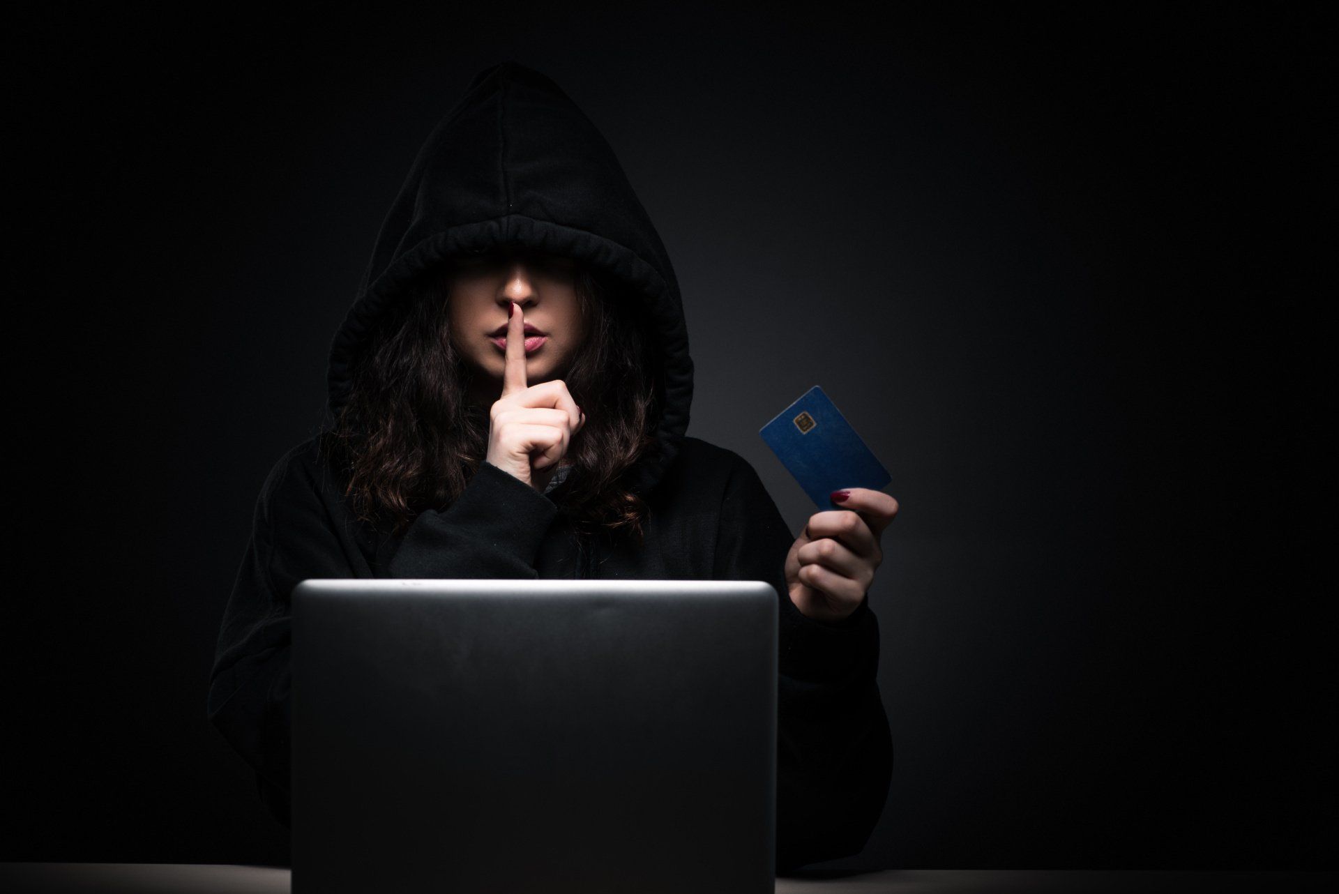 Hooded hacker holding credit card near laptop, representing stolen data and identity theft on dark web.