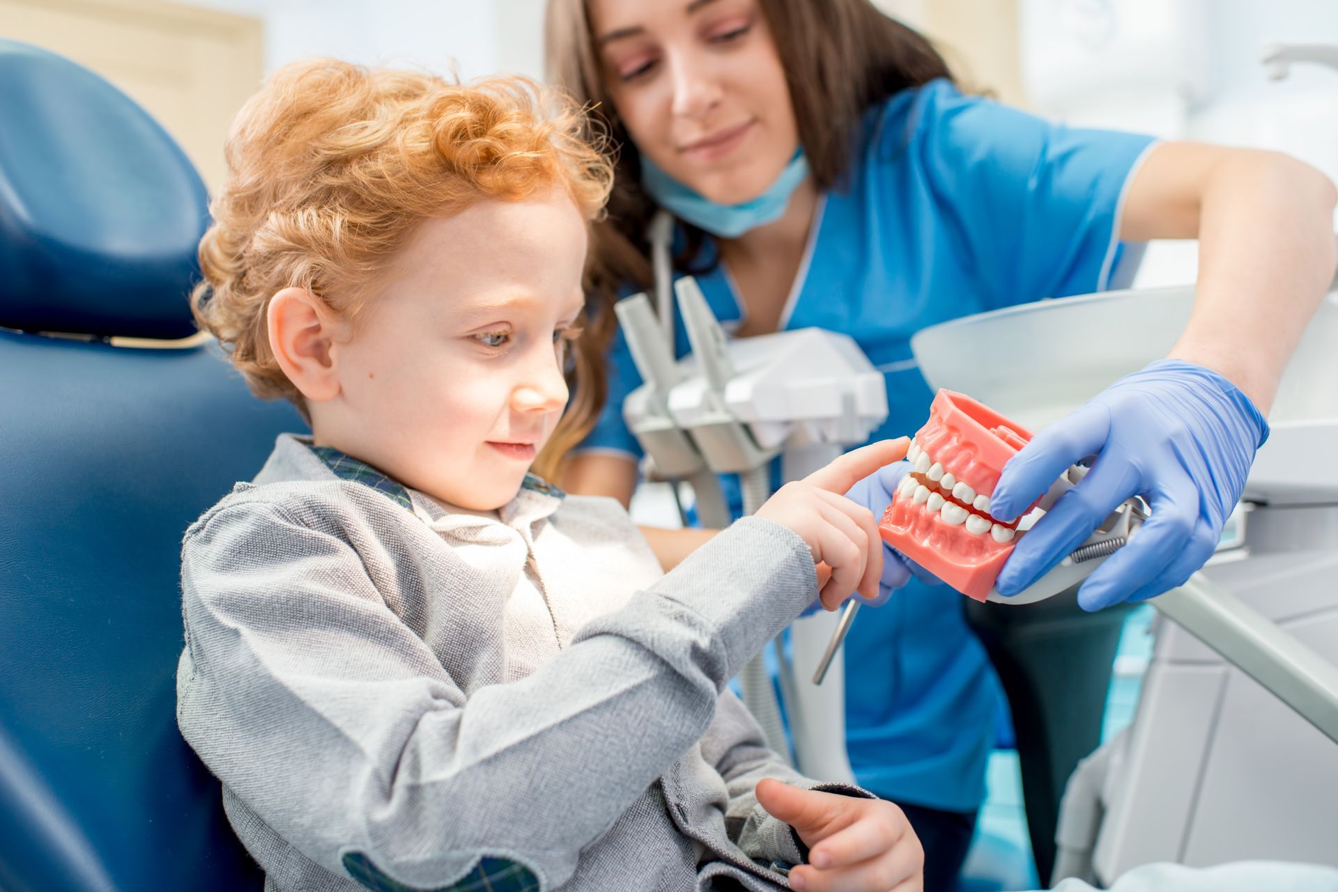 A dental professional wearing blue scrubs and gloves holds a model of teeth while a child points at it in a dental office.