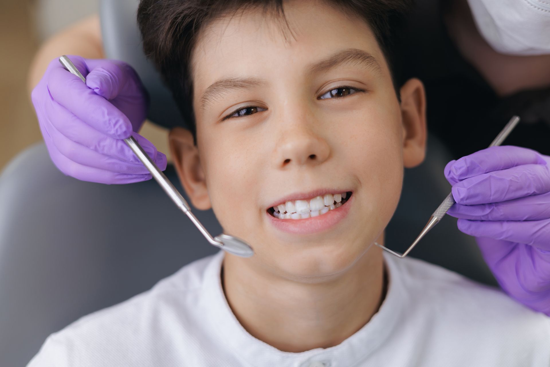 A person sits in a dental chair smiling while a provider in purple gloves holds a mirror and explorer tool.