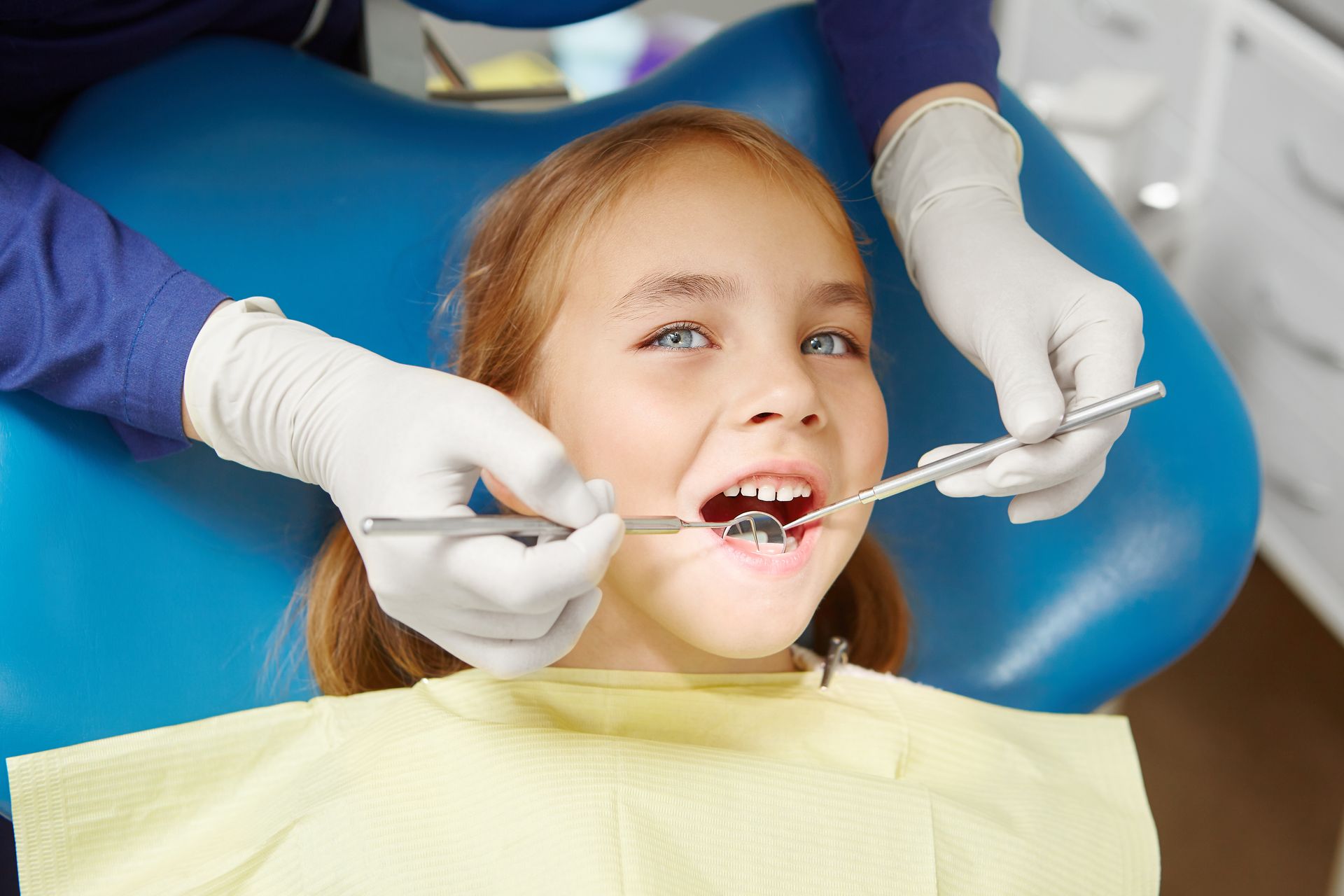 A dentist wearing white gloves uses a mirror and dental tool to examine a patient's teeth during a checkup.