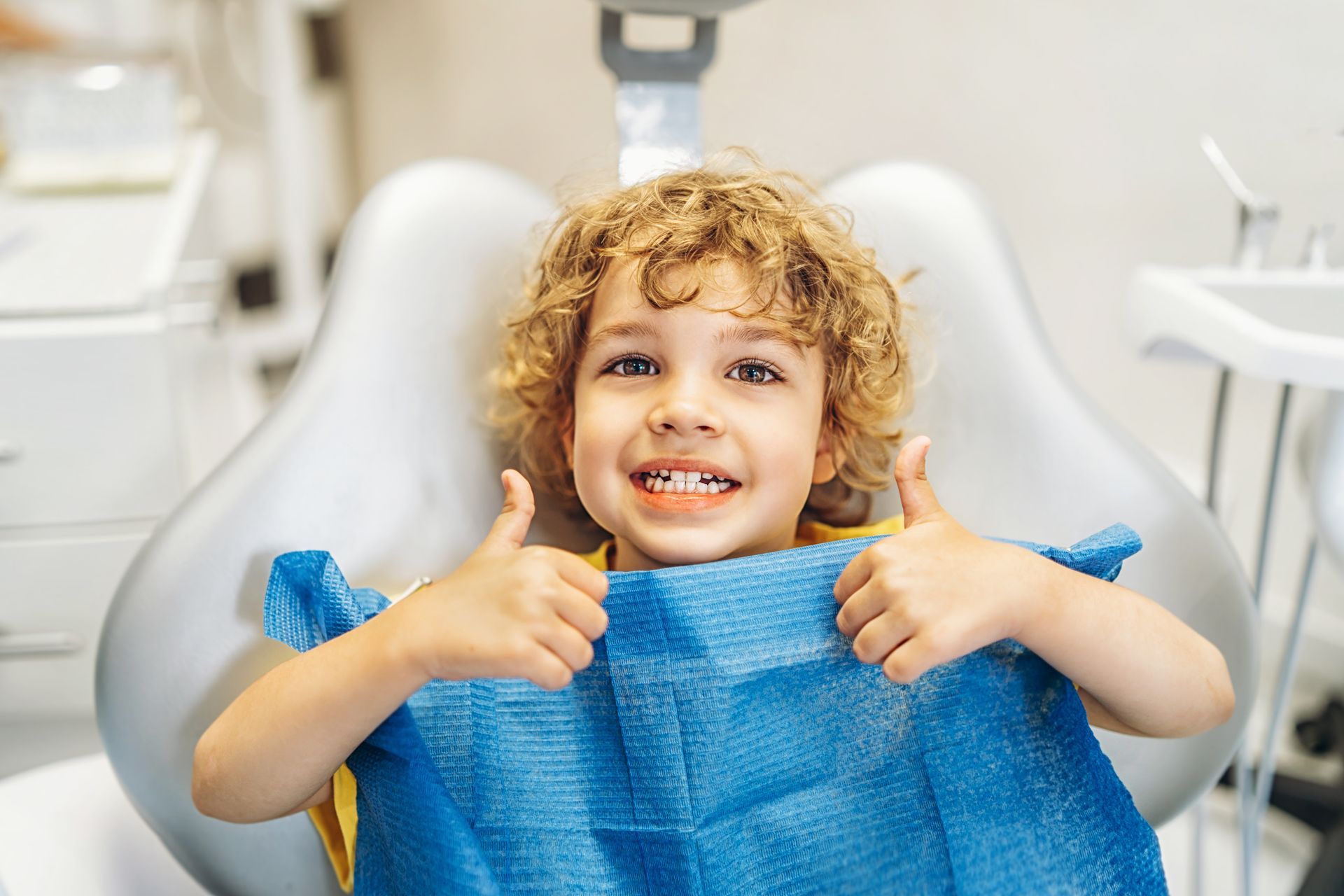 A smiling child in a dental chair giving a thumbs-up while wearing a blue bib.