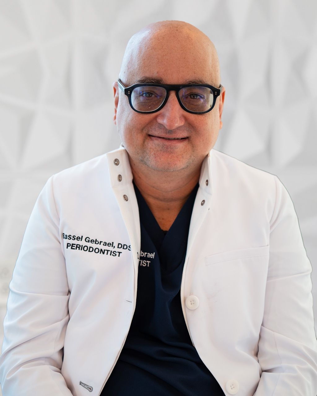 Man in white lab coat and glasses, smiling. Dentist with name tag visible.
