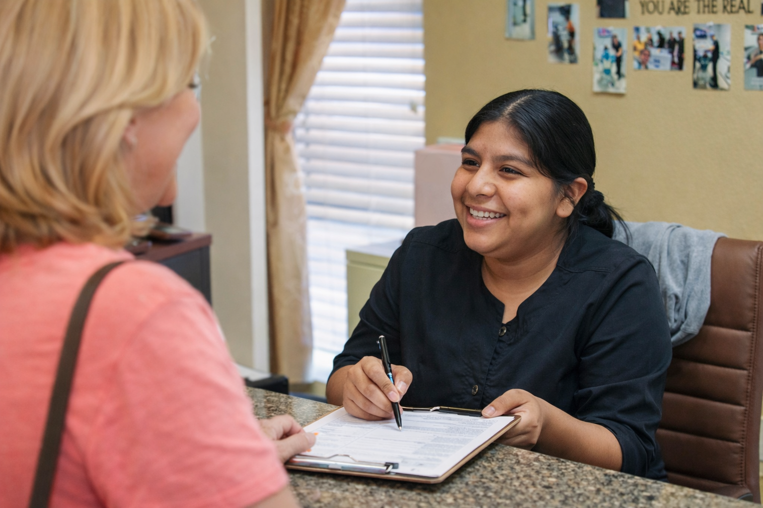 Two women talking at a table, one smiling and taking notes on a clipboard in an office.