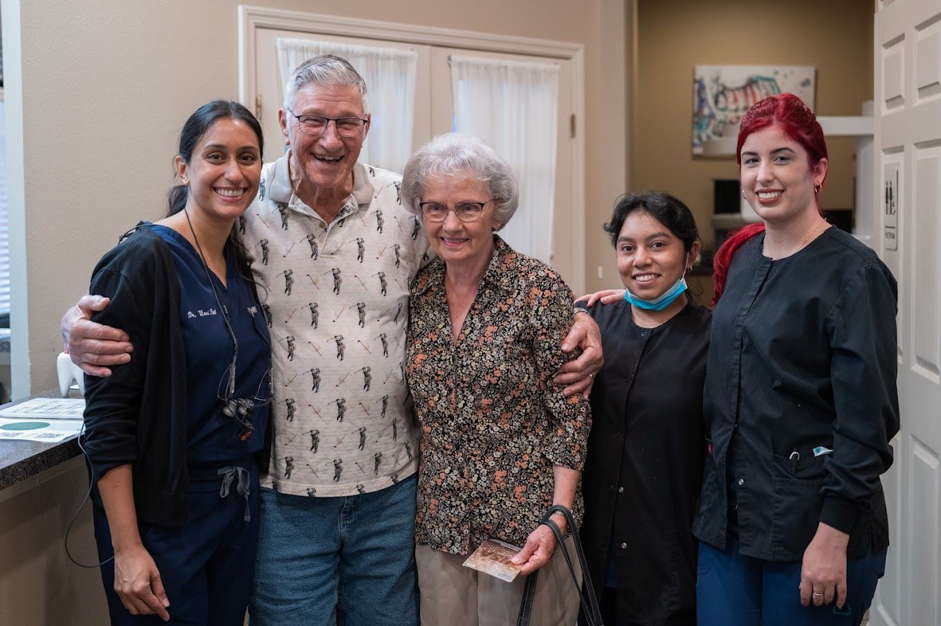 Five people smiling in a medical office; two older adults flanked by three dental staff.