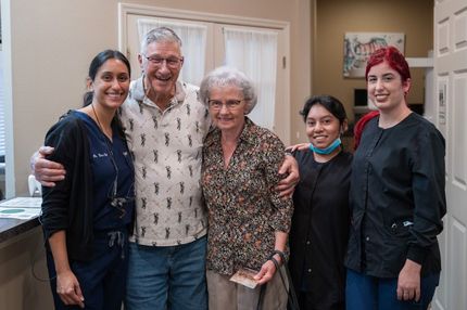Five people smiling in a medical office; two older adults flanked by three dental staff.