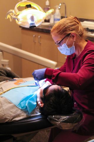 Dentist in maroon scrubs, mask, and glasses examining patient's teeth under a bright light.