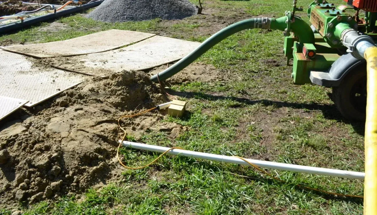 A green pump machine draws water through a hose into a white pipe located in a grassy construction site.