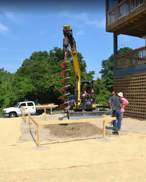 A yellow mini excavator with an auger attachment works inside a staked-out construction site near a house.