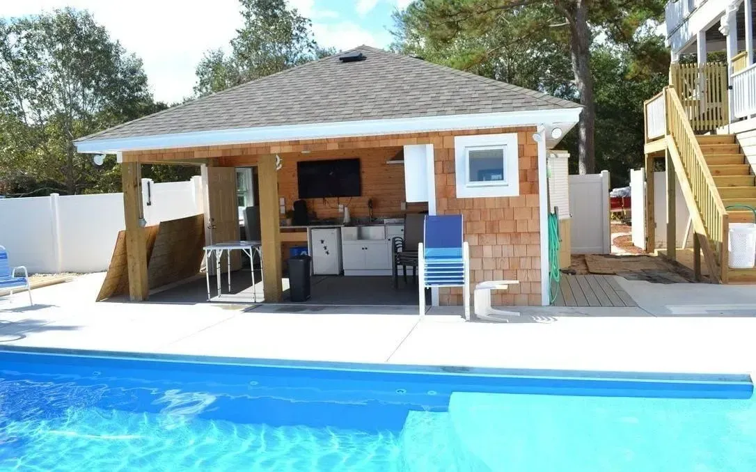 A pool cabana with an outdoor kitchen, TV, and seating beside a blue swimming pool on a sunny day.