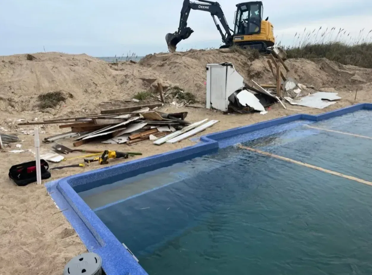 An excavator parked near a blue swimming pool on a sandy beach with scattered construction debris.