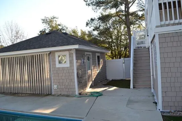 A swimming pool area beside a small, tan-shingled building with a grey roof and an outdoor staircase to a main house.