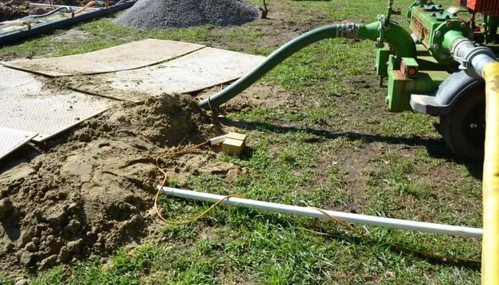 A green industrial pump sits on a grassy field, connected to a suction hose in a dirt pile and a yellow discharge hose.