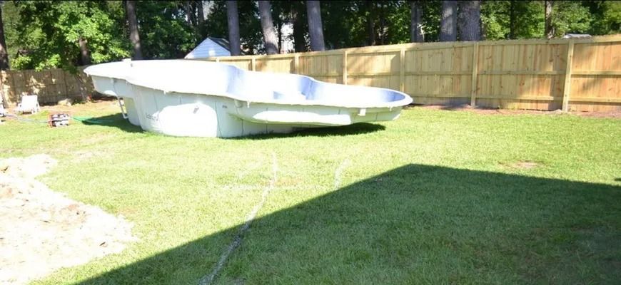 A white, empty fiberglass swimming pool shell sitting on a green residential lawn in front of a wooden fence.