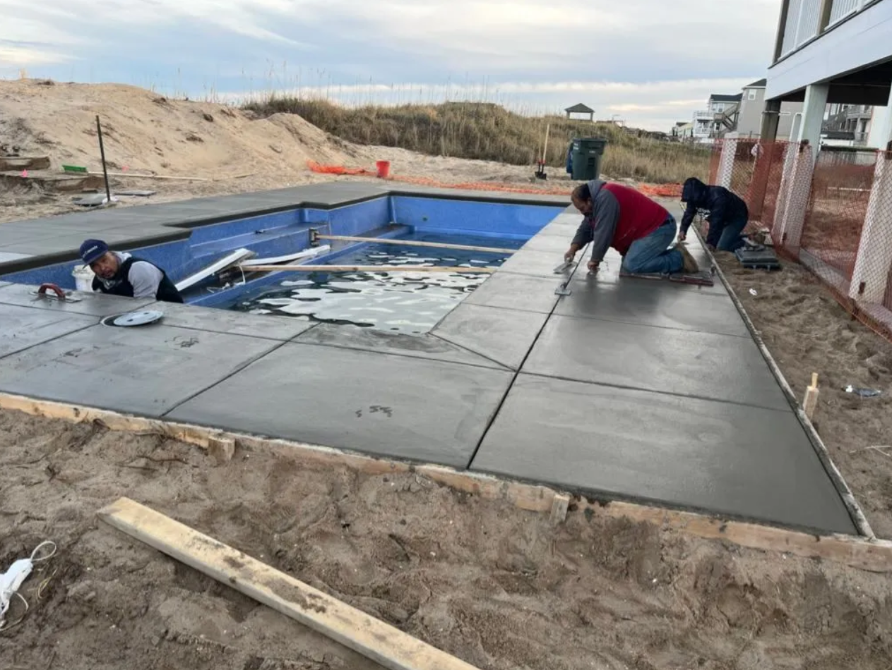 Three construction workers finish smoothing wet concrete around the edges of a backyard swimming pool under construction.