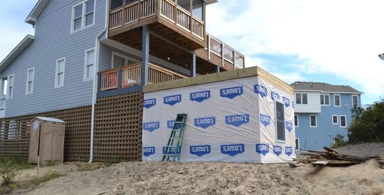 A blue house with a new addition covered in branded white house wrap, featuring a ladder leaning against the side.