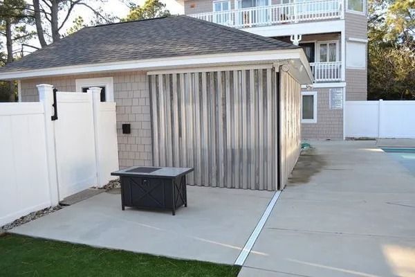 A small building with gray siding and metal hurricane shutters by a pool, fenced yard, and a patio fire pit.