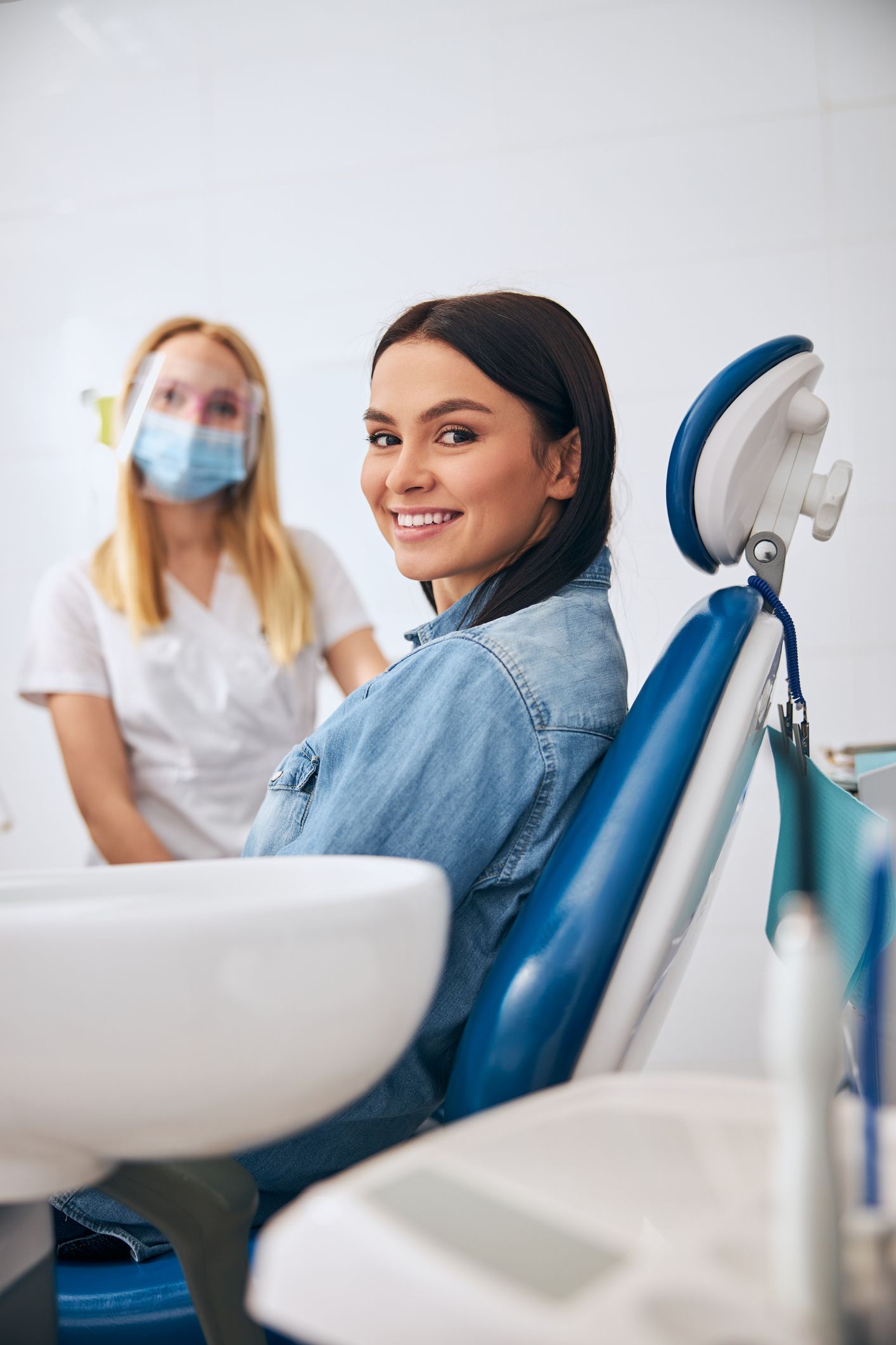 Smiling patient sitting in dental chair at modern dental centre with dentist in background.