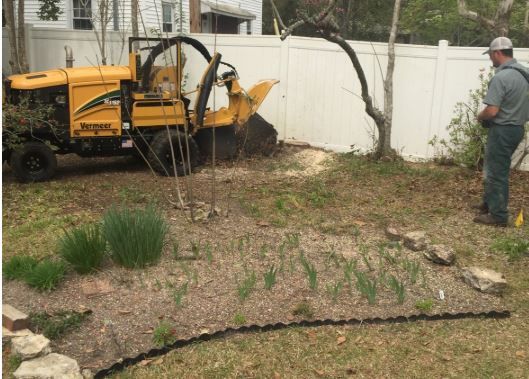 Yellow utility machine clearing debris by a white fence while a worker stands nearby in a yard