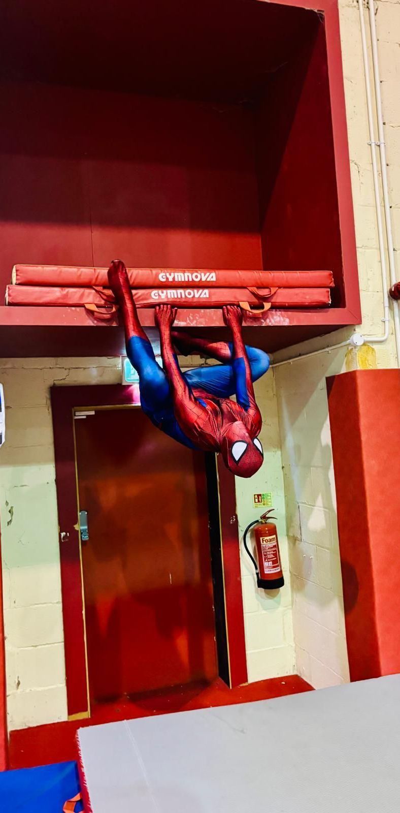 A spiderman is hanging upside down on a bar in a gym.