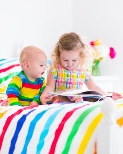 Two children sit on a bed with rainbow-striped bedding, looking at a book together.