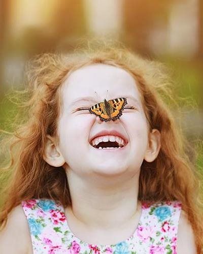 A red-haired child with a joyful expression and a butterfly resting on their nose, set against a blurred green background.