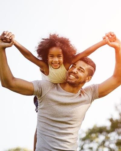 A person carrying a smiling child on their shoulders, both with arms outstretched against a bright, sunny background.
