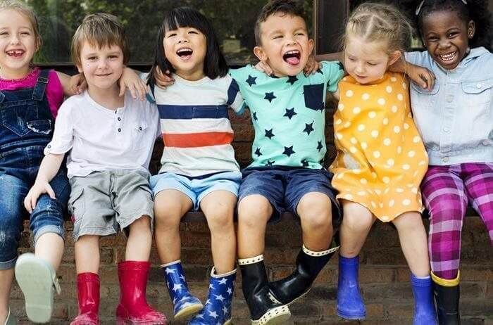Six children sit side-by-side on a ledge outdoors, all smiling and wearing colorful rubber rain boots.