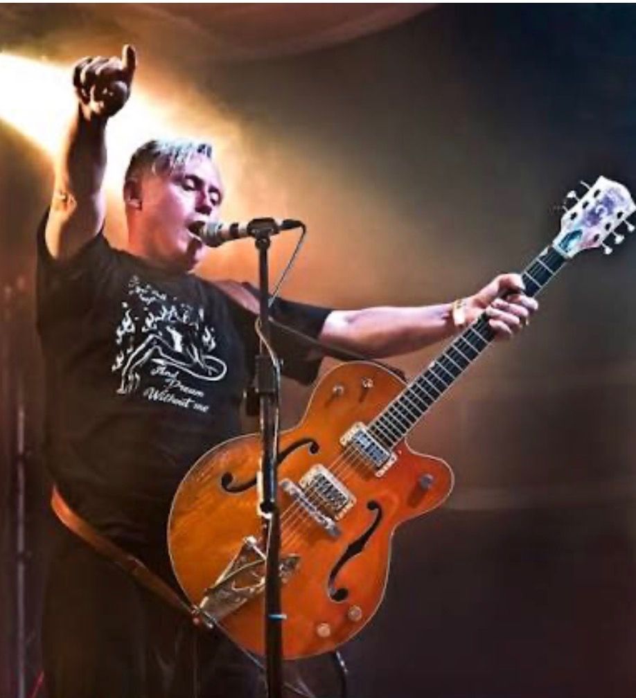 Man singing and playing a hollow-body electric guitar onstage, raising his arm.
