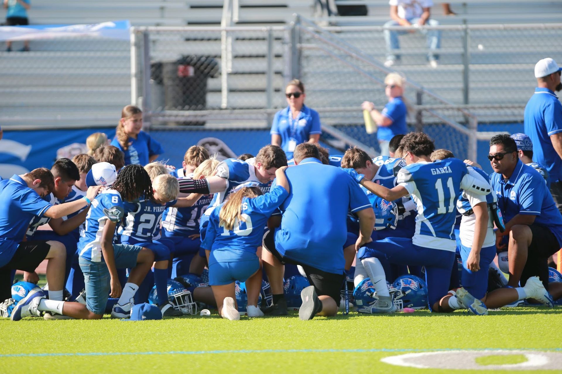 Football players huddle around a football, reaching hands together, blue sky backdrop.