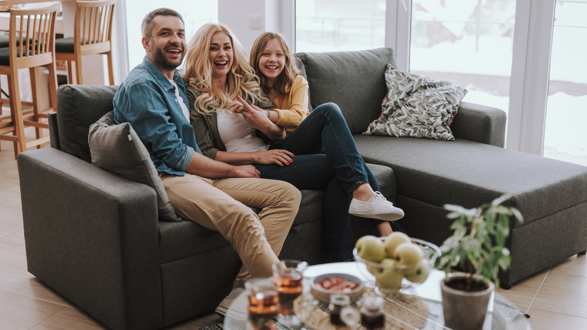 Family of three smiles while sitting on a gray sectional couch in a living room.