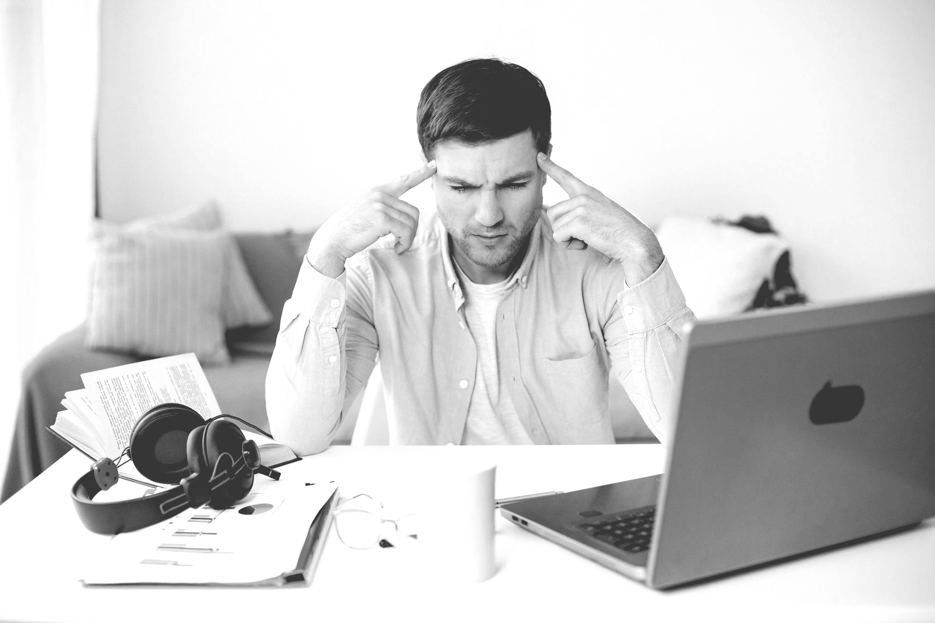 A person sitting at a desk with a laptop, looking stressed while pointing to their temples.