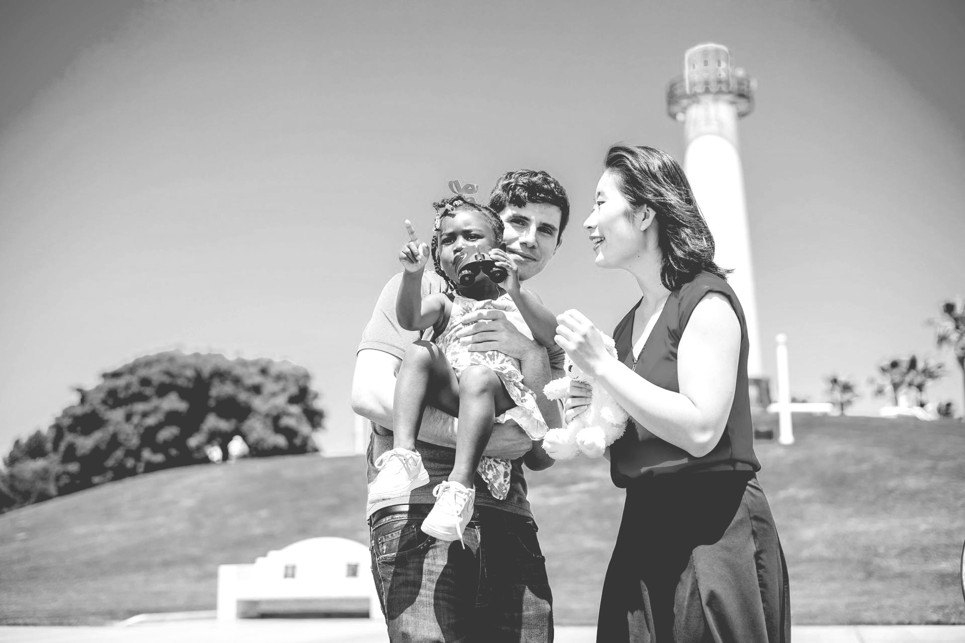 A family of three stands outside near a lighthouse on a sunny day, with the child pointing toward the distance.