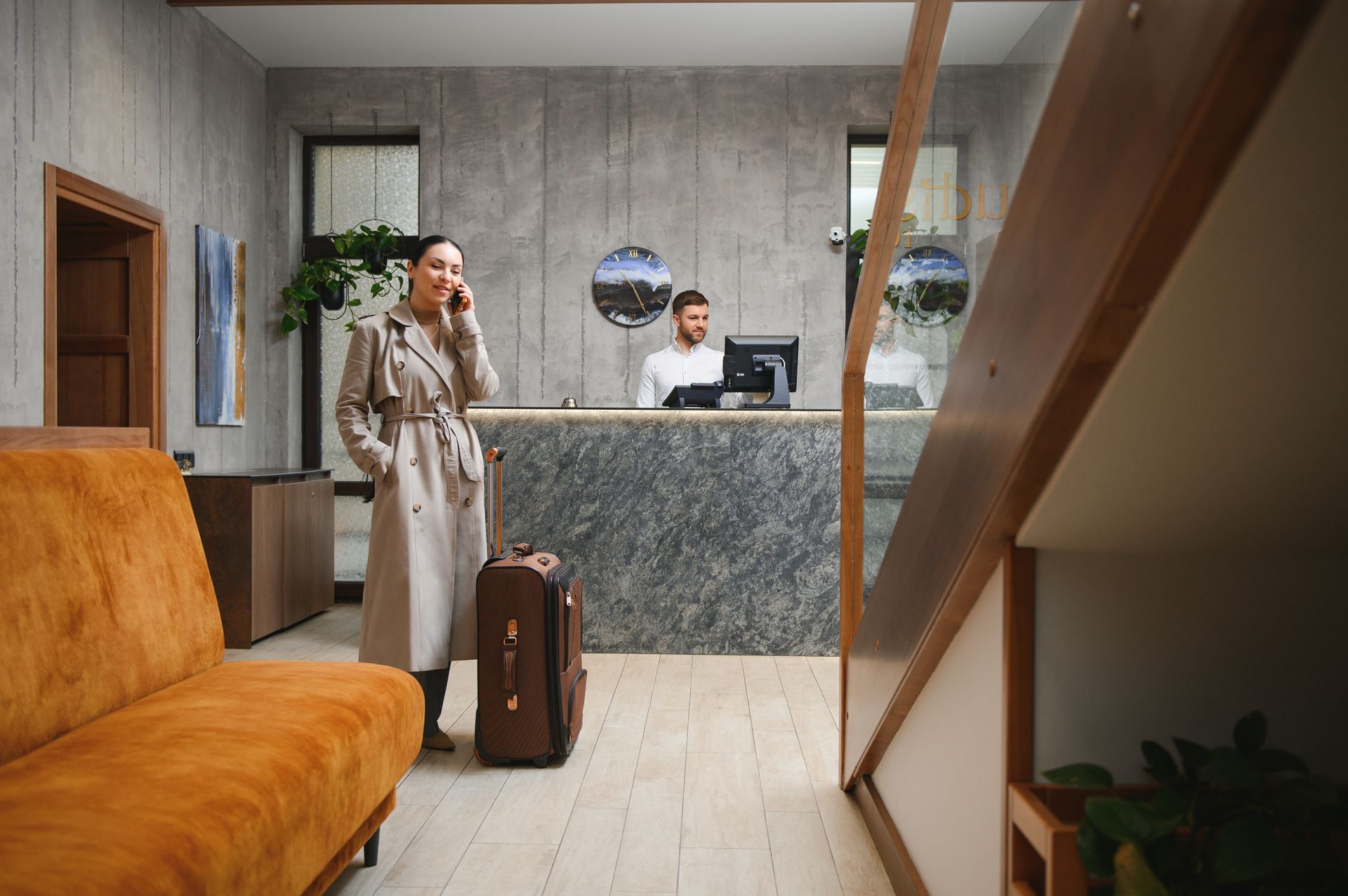 A person in a beige trench coat talks on a phone near their luggage in a hotel lobby with a receptionist in the background.