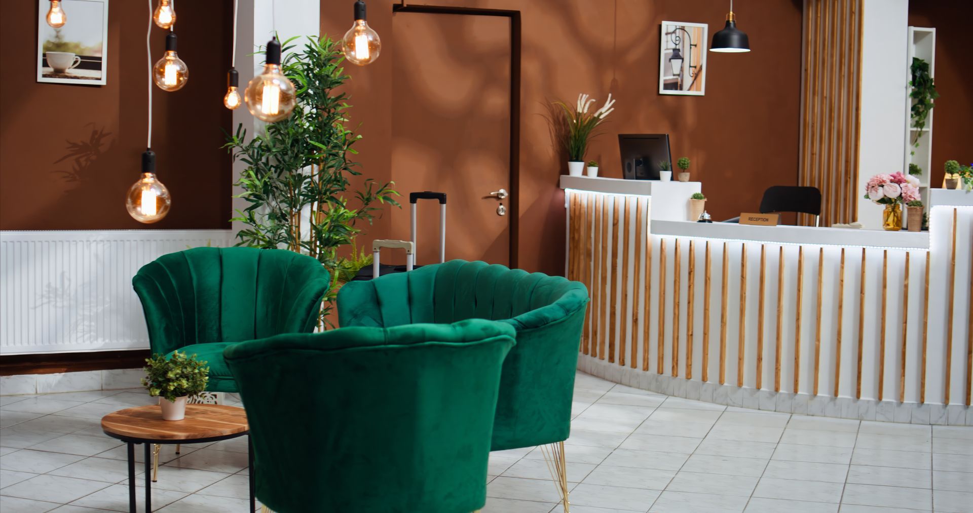 A modern hotel lobby featuring a white reception desk, green velvet armchairs, a wooden coffee table, and warm lighting.