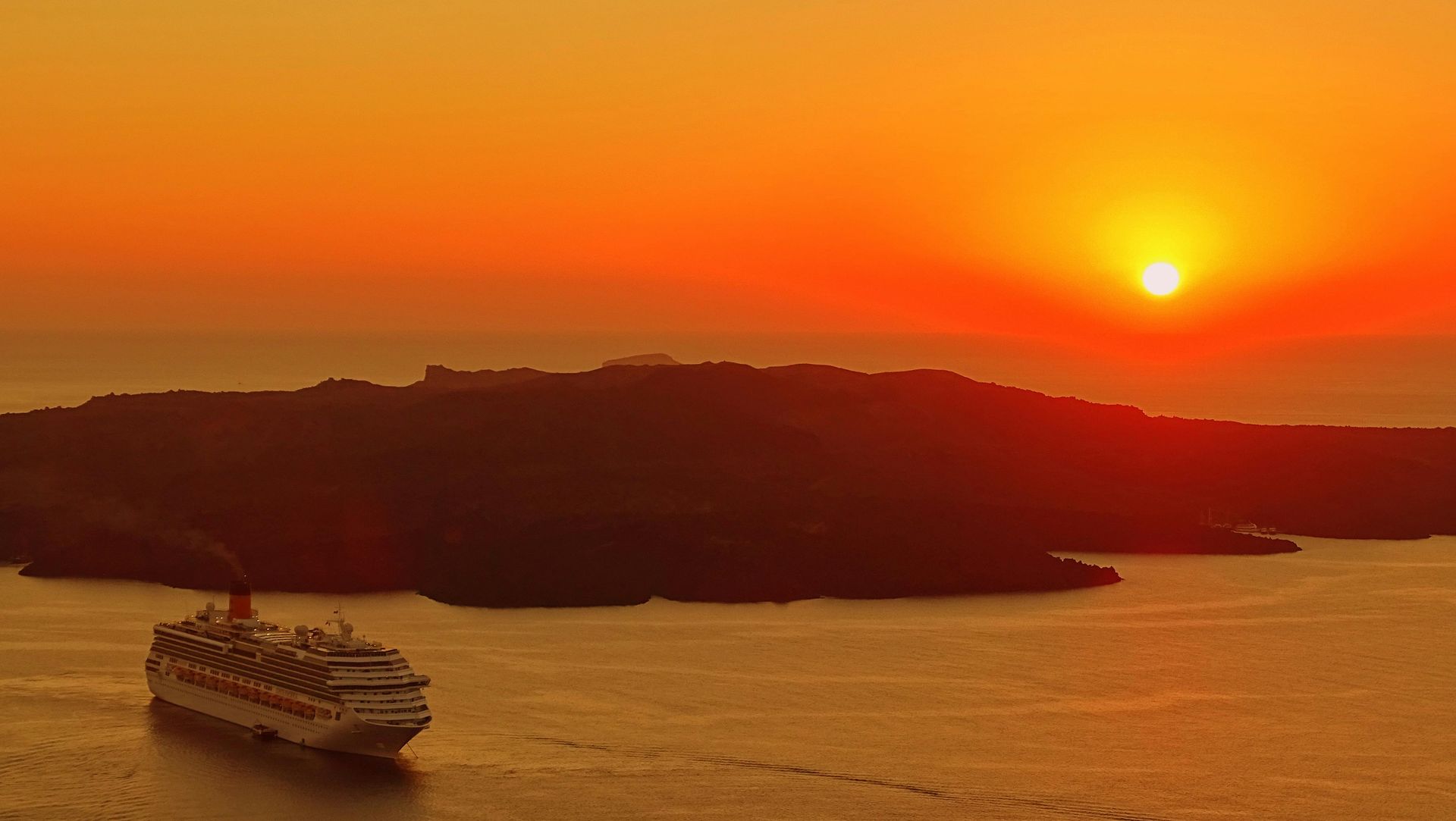 A cruise ship sails on dark water before a silhouetted island under a brilliant orange sunset.