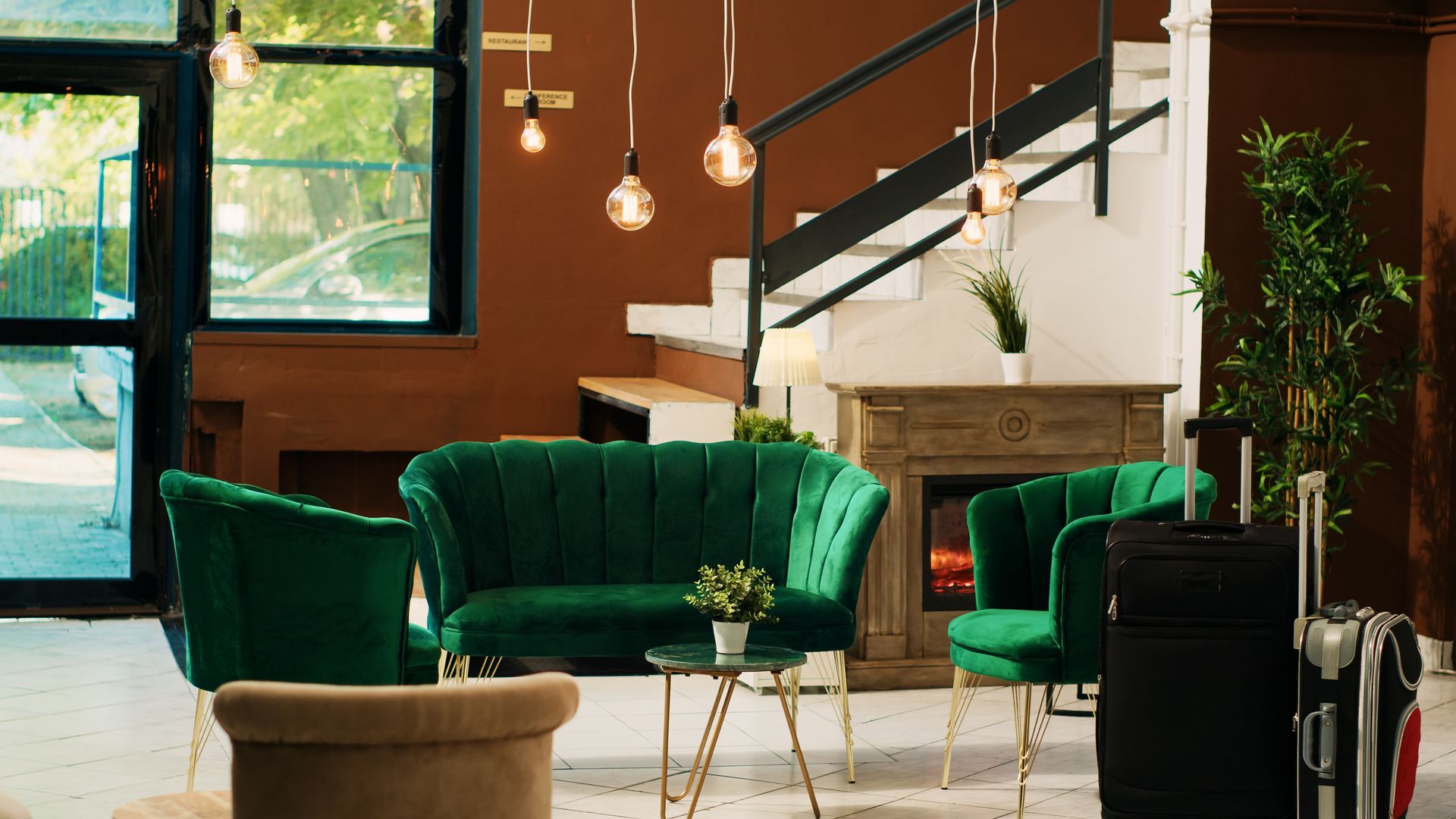 A modern hotel lobby featuring green velvet seating, a fireplace, hanging lights, and luggage.