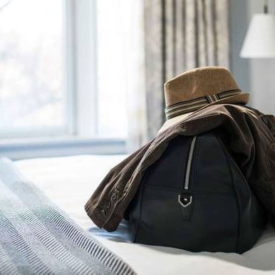 A tan fedora and brown jacket rest on a black duffel bag atop a white bed in a room with soft light.